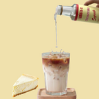 Cheesecake flavor syrup being poured into a glass of iced coffee with a slice of cheesecake on a yellow background.