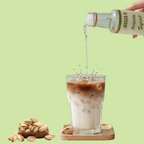 Pistachio syrup being poured into a glass of iced coffee with pistachios on a green background